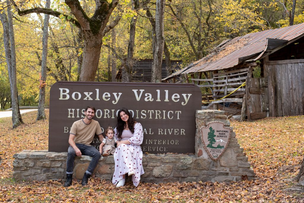 A couple and a small child sitting on a low stone wall with a sign behind them that says Boxley Valley Historic District.
