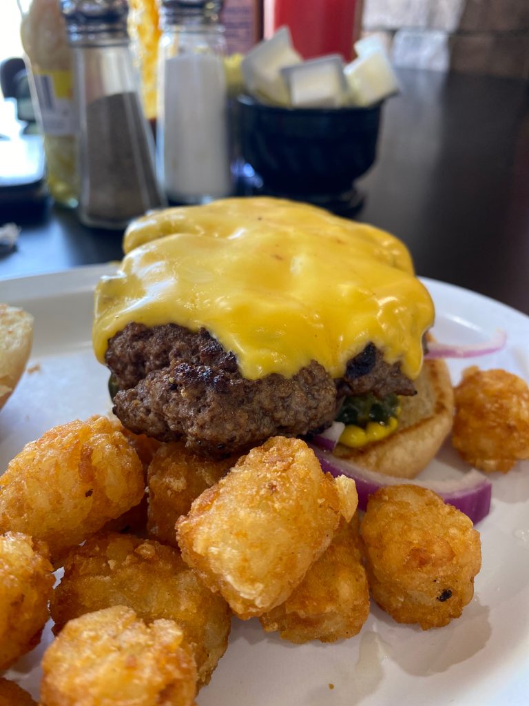 Close up of a juicy thick hamburger with American cheese on top and tater tots in the front. 