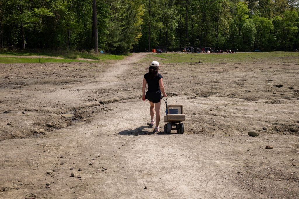 A girl walking in a field pulling a wagon with a bucket, a shovel, and some sifting items.