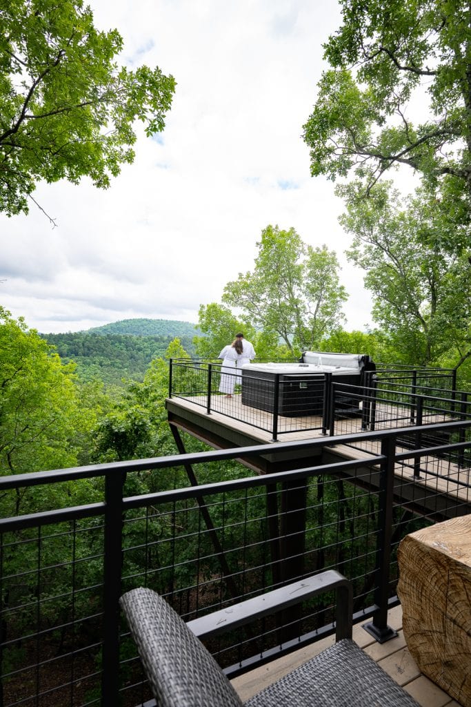 A young couple in robes looking out at a wooded Mountain View from a high deck with a hot tub.