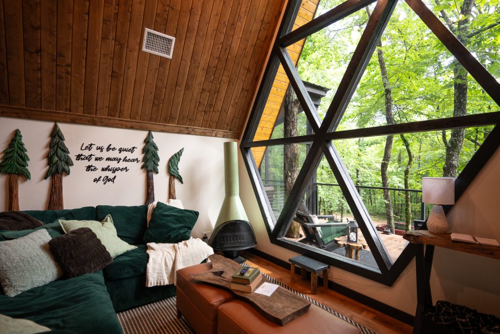 The interior of an A Frame cottage in Mena Arkansas. You can see through a huge window to the trees and there is a green velvet sofa. 