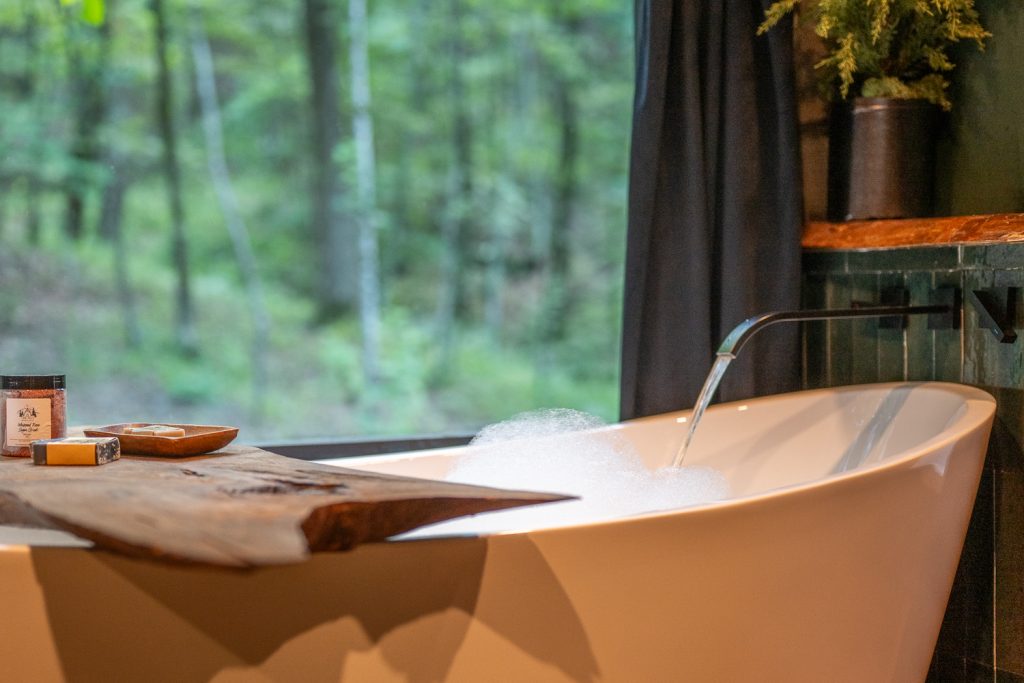 A bathtub with bubbles and a large window looking out into the woods.
