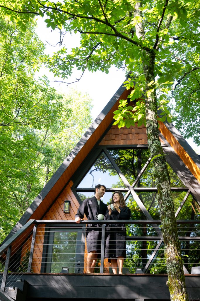A couple standing on the  deck of a house in the morning with their coffee. They are wearing black robes. 