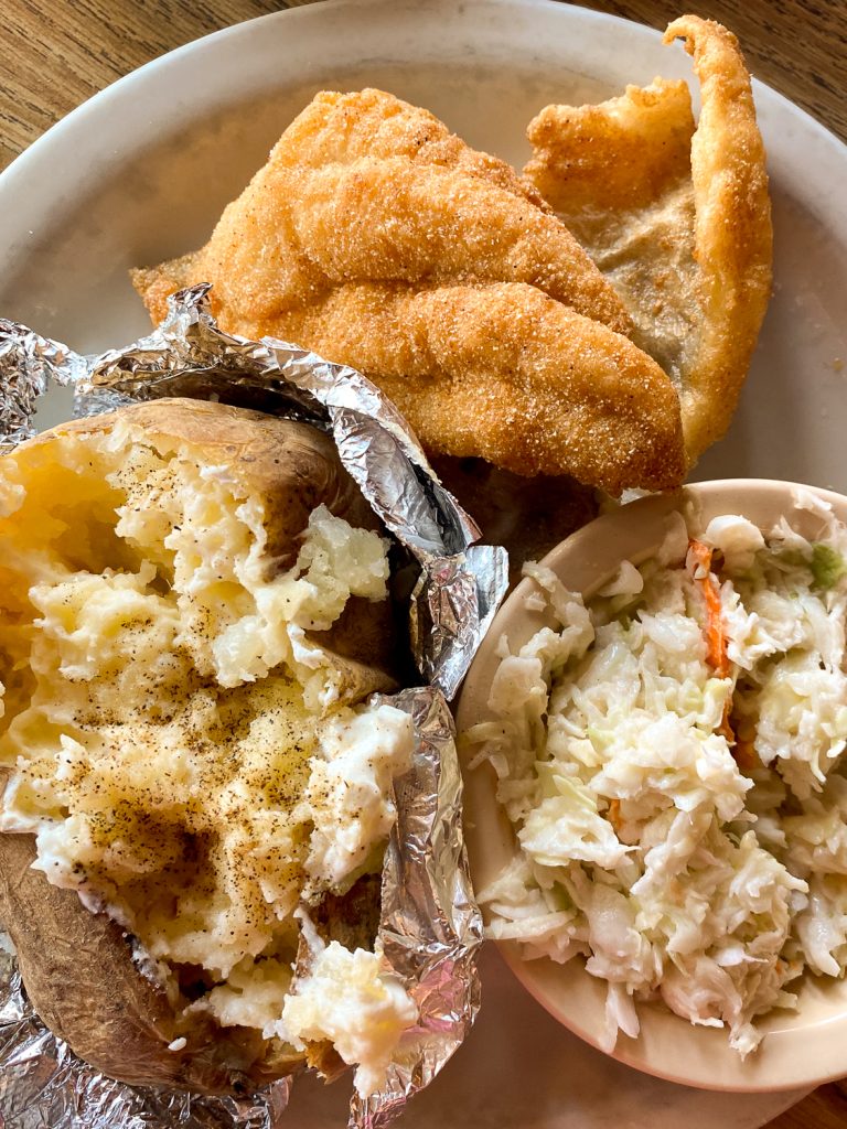 A photo taken from above of a plate with golden catfish, a baked potato, and coleslaw. 