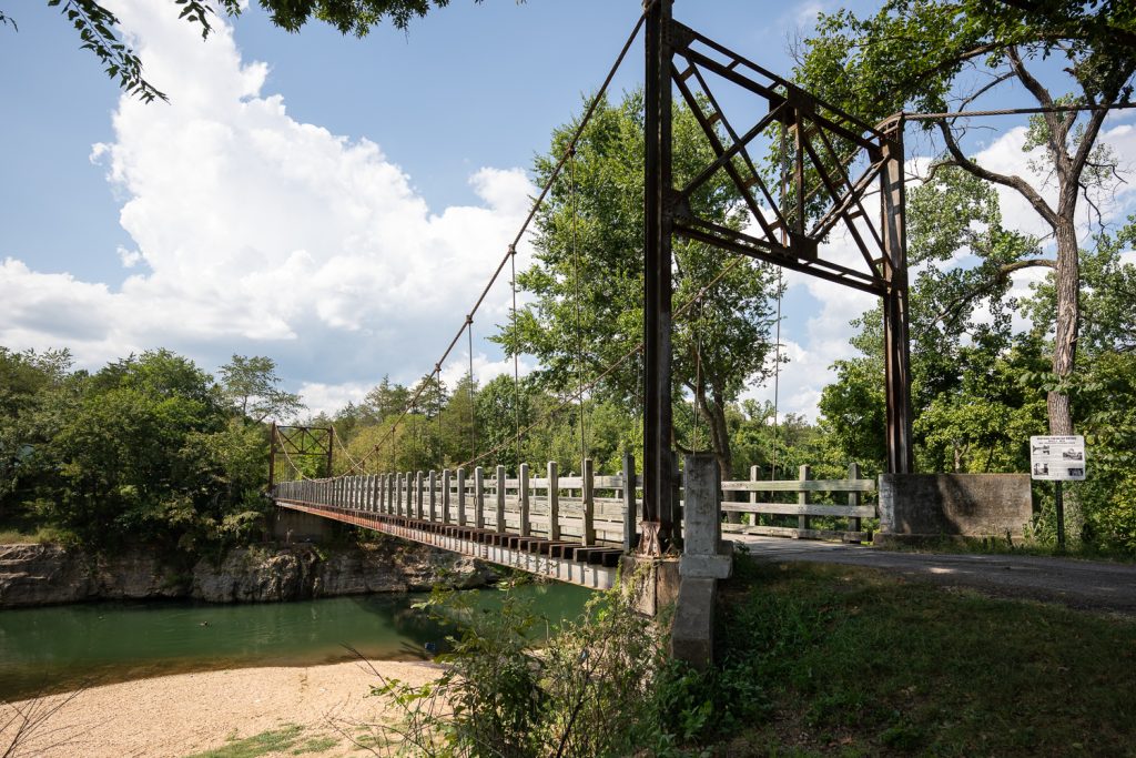 View of the Sylamore Creek Swinging Bridge in Arkansas. You can see the historic bridge from the side with the green waters of the creek below.