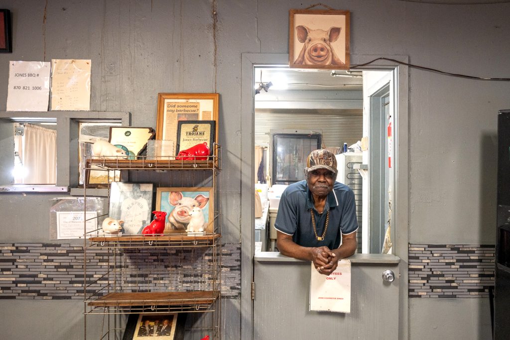 An older black man leaning on the half door of a window inside a barbecue restaurant. He is wearing a ball cap and a blue shirt. He is the owner of Jones Bar-B-Q Diner in Marinanna, Arkansas. 