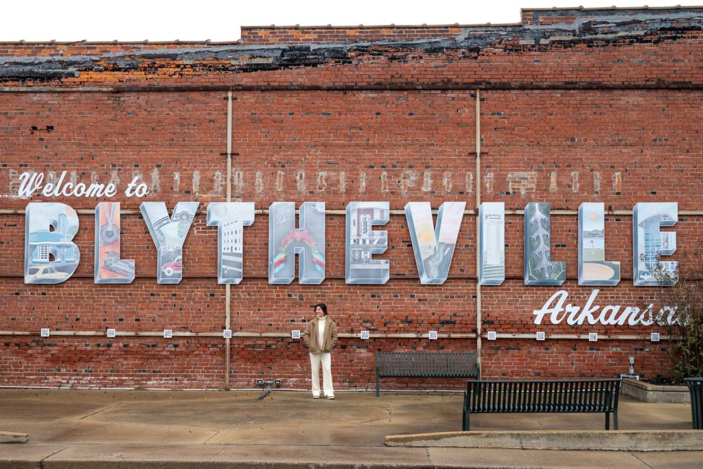 A sign on a red brick wall that say welcome to Blytheville, Arkansas. There is a young woman standing in front of the sign. This is part of a post about Christmas in Mississippi County Arkansas.