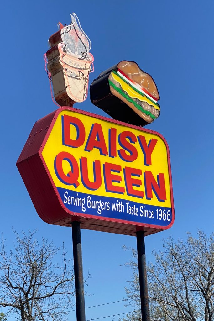 A sign silhouetted against a bright blue sky that has an image of a burger and ice cream cone on top and says Daisy Queen serving burgers with taste since 1966.