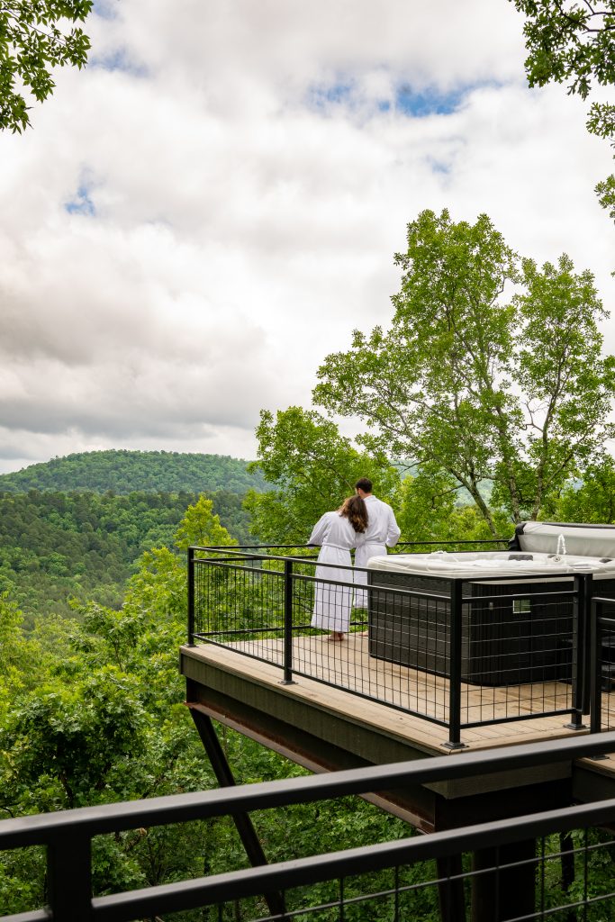 Couple standing on a deck looking out at the mountains. They are wearing white bathrobes and a hot tub is behind them.